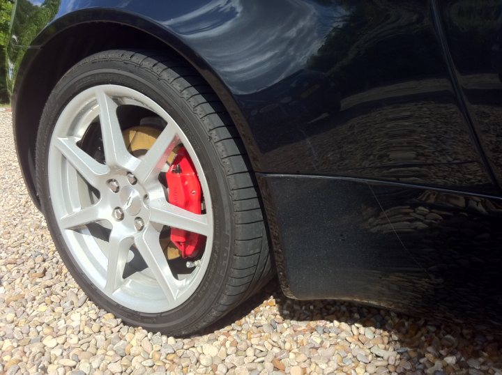 Pistonheads Film Grubby Protective - The image captures a close-up view of a black car's wheel and tire. The wheel looks modern and the tire indicates it's a non-summer, potentially all-season or touring tire given its tread pattern and sidewall. The car is parked on a gravel surface, a grey, pebble-like material providing a textured backdrop. The sunlight casts interplaying shadows and highlights across the scene, adding depth and dimension to the image. The view emphasizes the circular symmetry of the tire and the striking contrast between the black wheel and the lighter colors of the surface it's resting on.