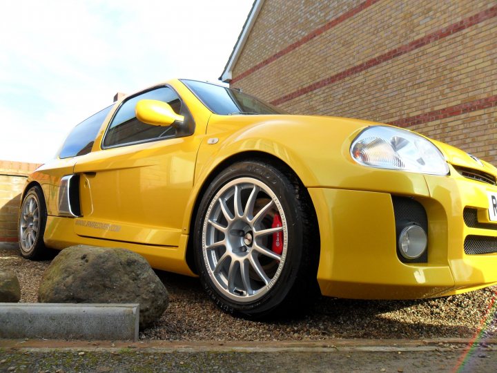 Speed Pistonheads Trials Brighton Sooner - The image features a vibrant yellow sports car parked against a red brick wall. The vehicle's front end is prominently visible, showing its sleek design and a pair of red reflector stripes on the silver hubcaps of the car. In the foreground, there are rocks, and on the left, a portion of a light post is visible. The sky appears clear, suggesting a sunny day.