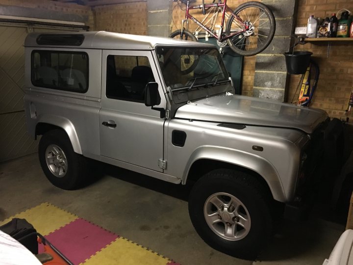 Pics of your offroaders... - Page 49 - Off Road - PistonHeads - The image features a silver-colored jeep parked indoors with a brick wall in the background. A bicycle is hanging on a hook above the jeep. A spotlight points at the front of the vehicle, illuminating the area around the wheels and bumper. The floor is tiled with various colors, including red, yellow, and black.