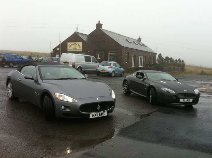 Pistonheads - In the image, two luxury cars are parked side by side on a wet road. One is a sleek black model and the other is a shiny silver vehicle. They are positioned in front of a small brick building, possibly a cafe or a pub, identifiable by a sign and a red awning. A few other cars are parked in the background, suggesting the place is not isolated. Despite the overcast grey sky and the relatively small number of vehicles, the scene conveys a sense of luxury and prestige.