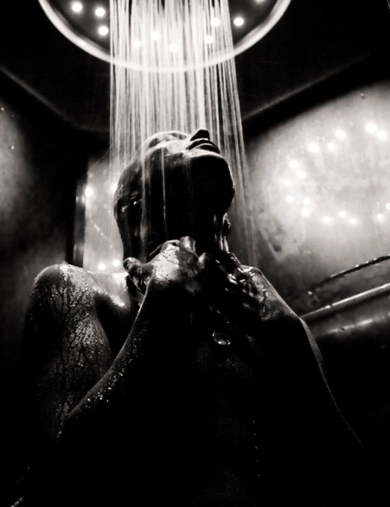 Girls Shower - The image portrays a person under an overhead shower. The light creates a dramatic effect, almost spotlighting the person, who seems to be in the midst of an intense shower. The background is dark and indistinct, making the shower the main focal point. The contrast between the person and the surrounding darkness emphasizes the soaking scenario. The overall atmosphere is one of solitude and perhaps melancholy.