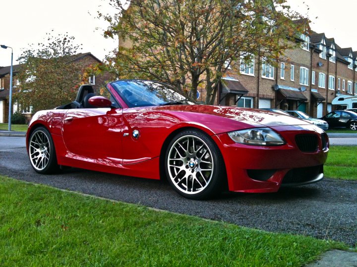 Imola Red Z4M Roadster - Page 3 - Readers' Cars - PistonHeads - The image depicts a vibrant red BMW sports car parked on a street. The car has a sleek design, with the top down, revealing its interior, which contrasts beautifully with the red exterior. It's positioned in front of a building, suggesting it might be parked near a residential or commercial area. In the background, there are trees and buildings, adding to the urban setting of the scene. The image captures the essence of luxury and style that the BMW sports car represents.