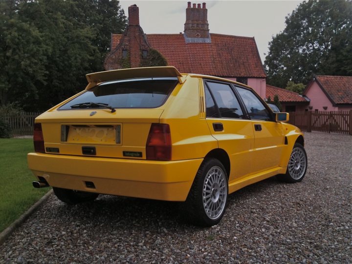 Endulgence Pistonheads - This image features a yellow Rover 200 compact car parked on a gravel driveway. The car is a two-door hatchback with a raised rear deck featuring a spoiler, which adds to its distinct design. Behind the Rover, there is a yellow three-story house with a distinctive chimney stack. The weather appears overcast, with the sky occupying the top third of the image. Additionally, there's a wooden fence to the right of the car, indicating a boundary for the property.