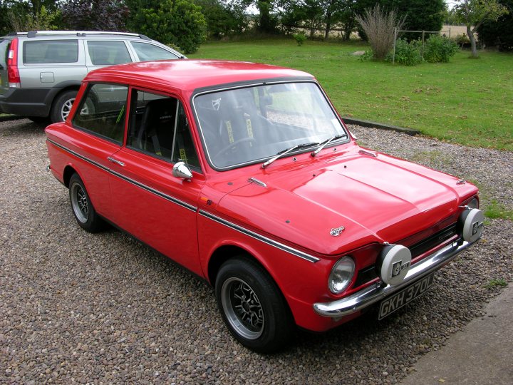 Pistonheads - The image shows a vibrant red vintage car parked on a gravel surface. The car has a shiny finish on its exterior, and its design features two round headlights and a rectangular grille, typical of classic cars. It has a distinctive black roof and black door caps, which contrast nicely with the red body. The car also has distinctive whitewall tires attached to silver-colored wire wheels. The gravel surface and the green field in the background suggest a rural or semi-rural location. This image also shows a car standing behind the vintage red car.