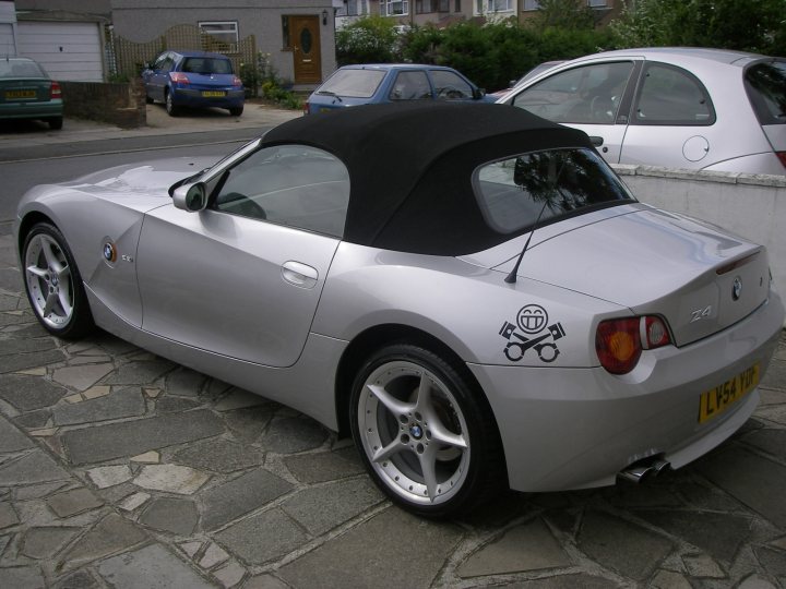 Pistonheads - The image features a convertible car with its top down, parked on a paved surface with a stone-tiled driveway. The car's paint is a shiny metallic silver color, reflecting the light. There is a sticker with a unique design on the rear, hinting at a particular symbol or logo that is not fully visible. The car's wheels are a silvery color matching the bodywork, and it stands out prominently against the backdrop of a residential area with vehicles parked along the street. The overall scene suggests a quiet, possibly suburban neighborhood with good maintenance of the vehicles.