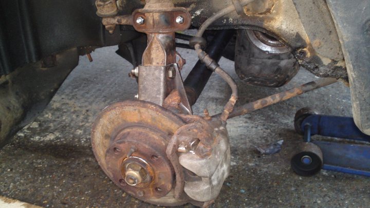 Project Peterborough - Page 3 - Alfa Romeo, Fiat & Lancia - PistonHeads - The image shows a close-up of a rusted brake drum with a rusted wheel stud visible. The drum is attached to a brake caliper with a disc brake pad, suggesting it's from a rear brake setup. The metallic brake pad is visible and looks worn. The image is taken on a concrete floor, which could be a workshop or garage due to the industrial appearance of the brake drum and other metal parts in the background. The overall condition of the brake drum and wheel stud indicates it's past its useful life and would require replacement and servicing.