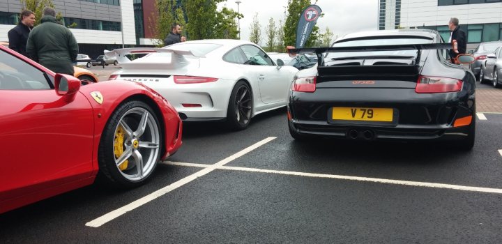 A car is parked on the side of the road - Pistonheads - The image captures a scene on a parking lot where several cars, including a red Ferrari and other white and black sports cars, are parked side by side. Multiple people are seen standing and talking next to the cars, suggesting they may be at a car event or a gathering of car enthusiasts. The ground beneath the cars appears to be wet, possibly due to recent rain. The setting is framed by a building in the background, adding context to the location of this event.