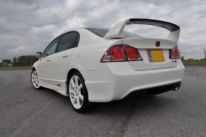 Import Type Pistonheads - The image shows a white sedan parked on a paved surface. The car has a distinctive rear spoiler and tail lights. There appear to be several red lights at the back of the vehicle. The background suggests it might be an outdoor area, possibly a parking lot, with what looks like chain-link fencing and an open space with structures on the horizon that could be buildings or structures.