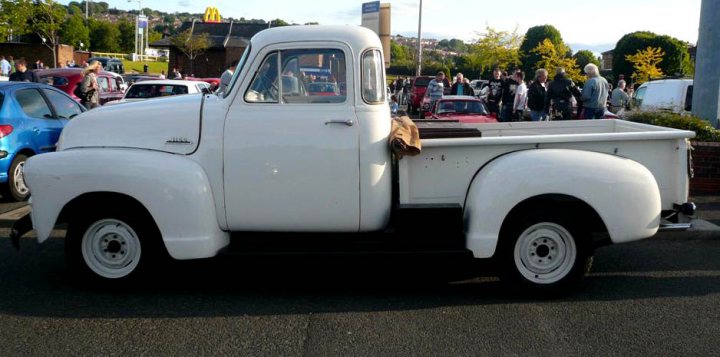 Pistonheads - The image features a white pickup truck parked on a tarmac, with people in the background who appear to be bystanders. The truck has a fully enclosed cargo area at the back, and there is a bag hanging in the back. It's an older model of a pickup truck. It's parked beside a fence and there is a lot of other memorabilia in the background, indicating possibly a car show. The people are scattered and seem to be enjoying the event or simply waiting for something.