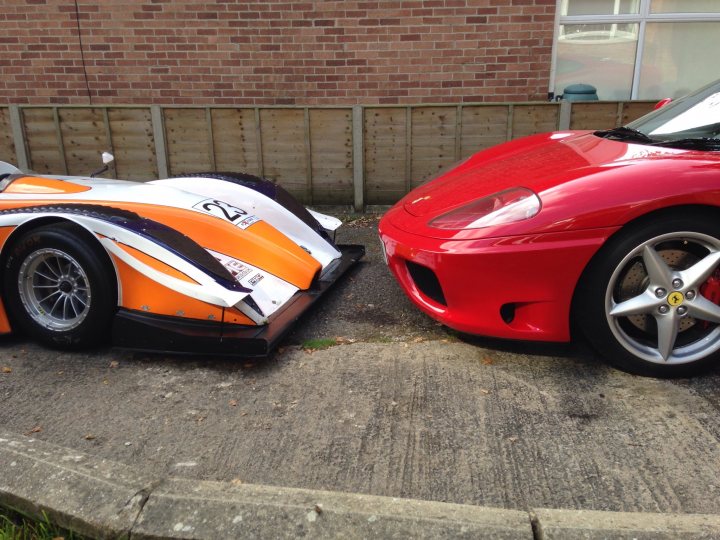 A red and black motorcycle parked in a parking lot - Pistonheads - The image shows two high-performance sports cars parked on a concrete surface next to a short wall. The car on the right is a bright red Ferrari, while the car on the left is white and black with an orange stripe. The cars feature distinctive design elements and showcase their aerodynamic bodywork and the small spoilers on their hoods. The atmosphere suggests the personal care taken in maintaining these high-end vehicles.