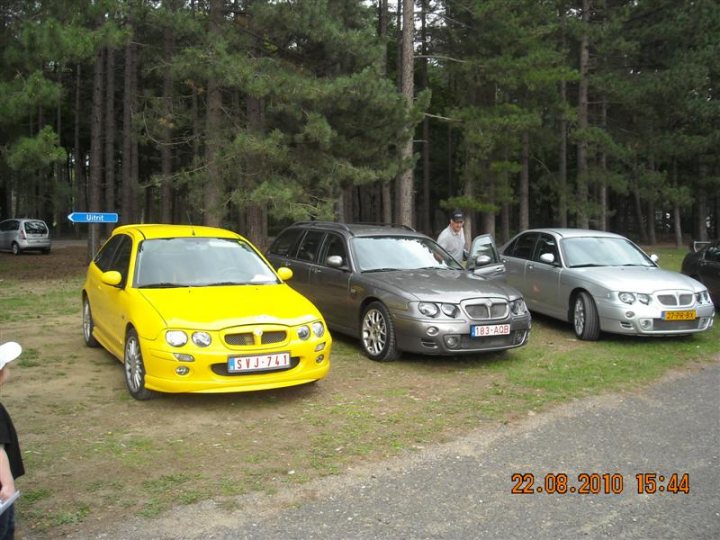 Pistonheads - The image shows a grassy area with a wooden trail on the left side, dotted with trees. Three cars of different colors and models are parked in a row by the side of the dirt road. From left to right, there's a vibrant yellow car that appears to be a hatchback, a silver car that looks like a luxury sedan, and another car with a patterned design that's more difficult to discern. The vehicles have European-style license plates. There's a person wearing a hat standing between the two front cars, and a small, white cap is visible near the far left of the image. The timestamp in the upper right corner indicates the photo was taken on the 22nd of August, 2010 at 4:44.