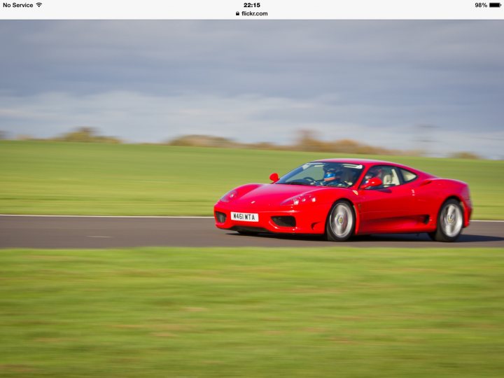 A red and white car with a red bow - Pistonheads - The image shows a red sports car driving on a road with grassy embankments on either side. The car is captured in motion, blurred to emphasize its speed. The time stamp in the corner reads "10:55", and there is a website URL, "flick.com," also present in the image. The setting appears to be wide-open country or a rural road, with an expansive view of the grassy landscape. The car emits a sense of motion and dynamism, indicative of a sporty or exotic vehicle for sale.