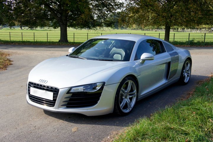 Audi RS / S / R8 picture thread! - Page 1 - Audi, VW, Seat & Skoda - PistonHeads - The image showcases a beautifully detailed silver Audi R8, parked on a driveway with lush green grass on both sides. The car's sleek design is accentuated by the large black grille that houses the distinctive Audi emblem. The rear view and side mirrors, also in black, add a touch of elegance to the car's overall aesthetic. The dramatic skies suggest it might be late afternoon or early morning, with the soft, diffused light casting a warm glow on the car.