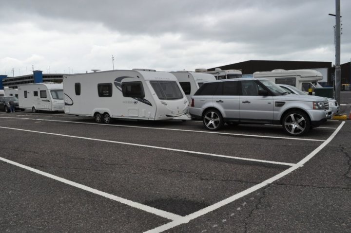 A white van parked in front of a building - Pistonheads - The image shows a parking lot with a variety of recreational vehicles. There is a trailer that has been attached to a vehicle, possibly a trailer for a boat. A large station wagon is parked next to the trailer and is also towing a camper. Adjacent to these vehicles, there is a fifth wheel camper parked across the parking lot. A few other vehicles and trailers are dispersed in the background. There are no people visible in the image. The overall atmosphere suggests a trip or journey involving camping or outdoor recreation.