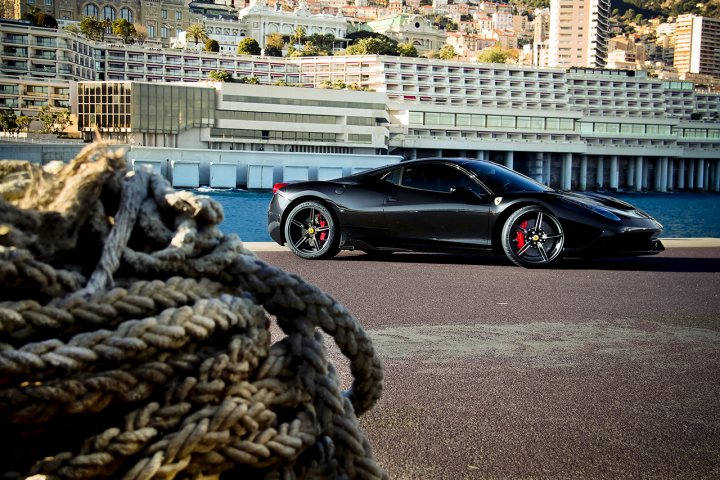 458 Speciale - Page 1 - Ferrari V8 - PistonHeads - In the image, a black sports car is the main focus, parked on a paved surface with a frayed rope leaning against it. The car is positioned on a waterfront, showcasing the vehicle's sleek design amidst the urban backdrop. The car's red rims provide a striking contrast against the backdrop. The city beach appears serene and quiet. In the background, there's a large building with a distinct architectural design, adding an interesting contrast to the natural waterfront scene. The image captures a peaceful moment, where the fast-paced urban life coexists with the tranquility of nature.