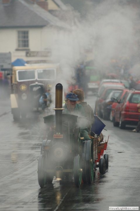 Classic tractors - Page 3 - Classic Cars and Yesterday's Heroes - PistonHeads - The image depicts a vibrant scene on a wet, misty street. A steam locomotive, with its additional seat in the back, is being pulled by one man from the back and another man from the front. The locomotive is moving down the street, amidst other vehicles. The street is populated with various parked cars and there's a person in the background holding an umbrella. The overall atmosphere of the photograph is nostalgic and tranquil.