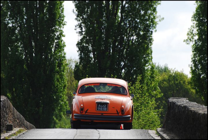 The Tour Auto came to town - Page 1 - Classic Cars and Yesterday's Heroes - PistonHeads - This image captures a serene scene on a country road. An old, orange Volkswagen Beetle is driving away from the viewer, its body and license plate clearly visible. The road is lined with tall trees, their green leaves forming a canopy overhead. The trees, which extend to the edges of the image, provide a sense of enclosure and add a touch of wilderness to the scene. To the right of the road, a stone wall can be seen, further emphasizing the rural setting. The entire scene conveys a sense of peace and tranquility.