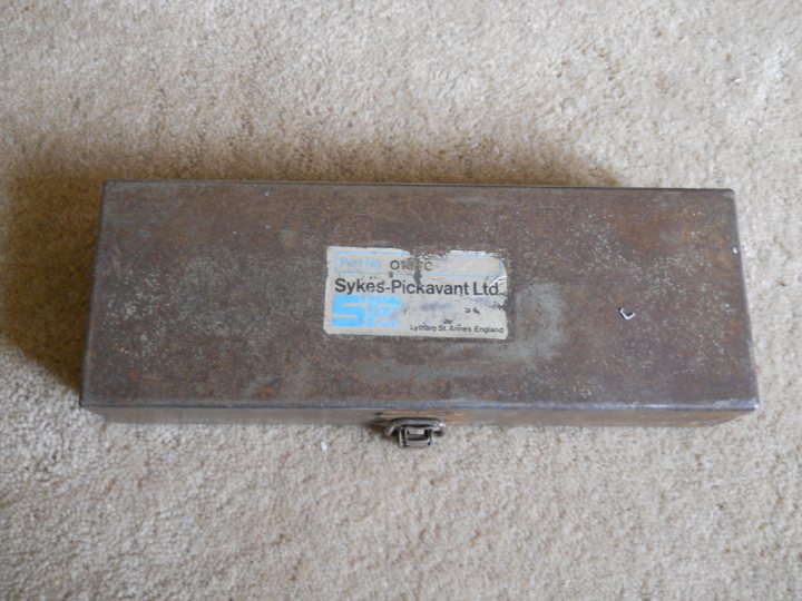 Name this tool! - Page 1 - Home Mechanics - PistonHeads - The image shows a closed mahogany wooden box lying on its side on a soft surface that appears to be a rug or a mat. The box is small, box-like with rounded corners, and has a metallic latch and lock mechanism visible on one of its long sides. There's a label attached to it, with some text and numbers, but the details are not clear. The background is mostly beige, contrasting with the darker tones of the box. The focus of the image is on the item itself, which is the main subject. The overall style of the photograph is straightforward, capturing the object from a perspective that allows the viewer to see its features, such as the type of material and locking mechanism.