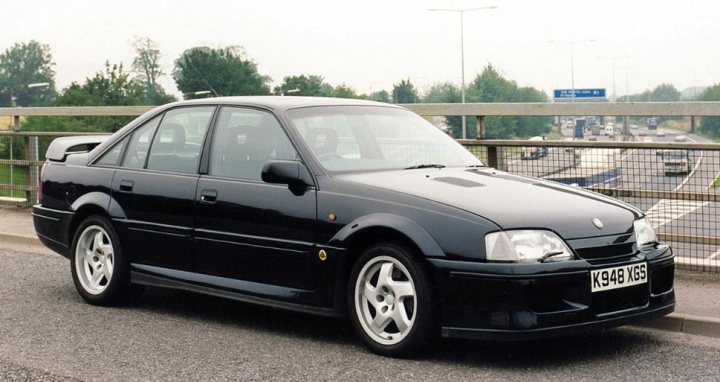 Pictures of decently Modified cars [Vol. 2] - Page 300 - General Gassing - PistonHeads - The image depicts a two-door black car parked beside a gray, empty road. The car's license plate reads "K84 KX3," with a noticeable dent on the driver's side door. The side of the car features a stainless steel strip along the lower side panel, which also has a crest logo visible on the front side panel. In the background, there's a white overpass with a blue road sign, indicating a highway that appears to be elevated above the surrounding area. The sky is gray and overcast, suggesting an overcast or cloudy day.