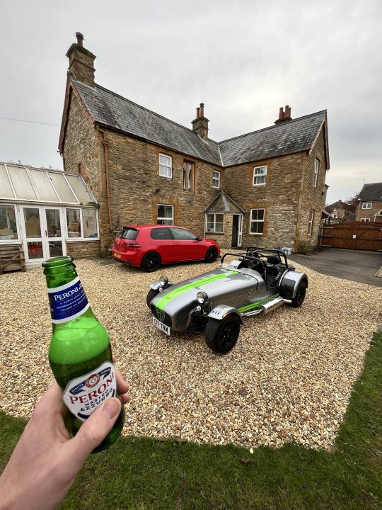 The image shows a person holding up a bottle of beer, with the focus on the hand and bottle. In the background, there is a residential house with a gray roof and a white brick chimney. Parked in front of the house are two cars: one green and another white. To the right, there's a motorcycle parked on gravel. The driveway appears to be made of small stones or pebbles. The setting suggests a quiet residential area during what might be late afternoon or evening.