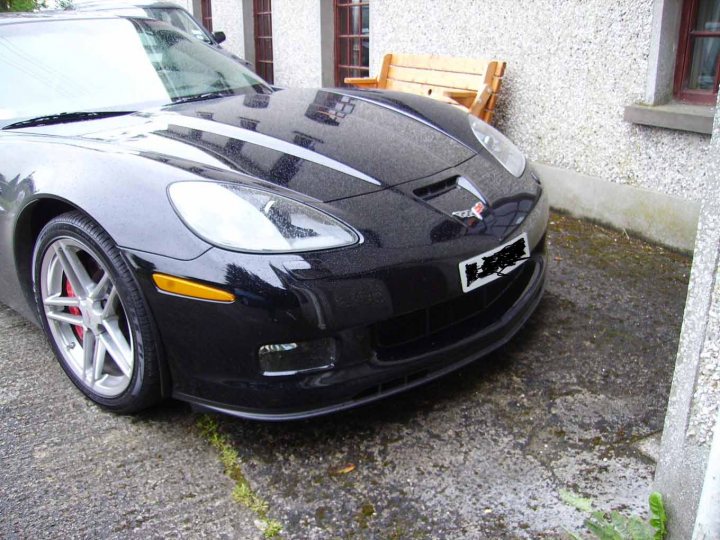 Pistonheads - The image shows a black sports car parked on a driveway alongside a building. The car features a sleek design with a prominent air intake at the front, and it is positioned at an angle that gives a view of its front and side profile. To the right of the car, there is a wooden bench, suggesting an outdoor seating area. The setting appears to be a residential or semi-urban area with a mix of textures visible on the road and ground.