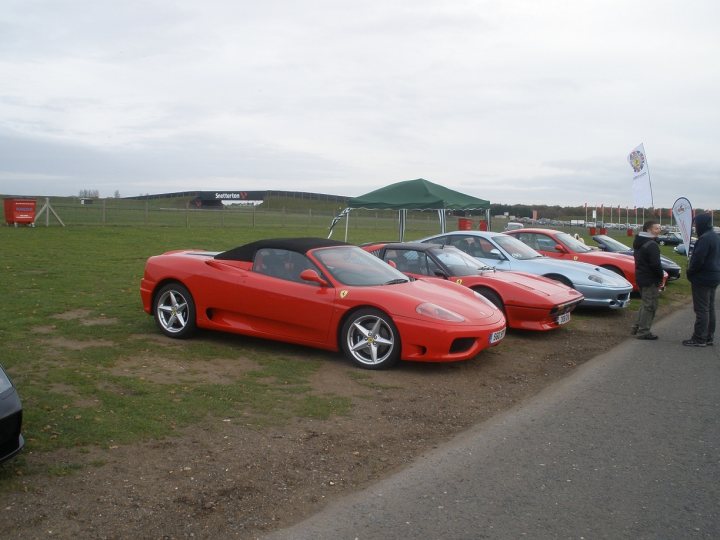 Thank you. - Page 1 - East Anglia - PistonHeads - The image depicts a gathering of people and cars on a grass lot. There are two red sports cars parked side by side. The cars are sleek and polished, clearly the focal point of the scene. In the background, several other cars are parked, and behind them, there are white flags fluttering in the breeze. The sky is overcast, adding a dramatic effect to the cool dusk setting. The people standing near the cars appear to be engaged in conversation or admiring the cars, creating a lively and social atmosphere.