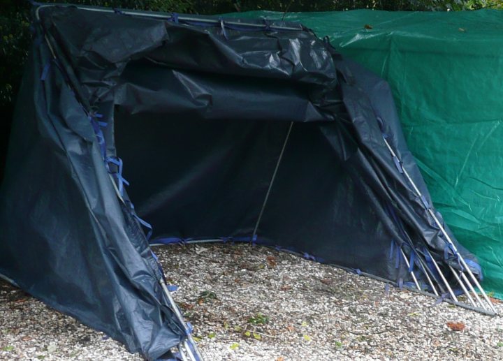 Carcoon Pistonheads Outdoor - The image shows a dark blue tent with a partially open door, set up on a gravel surface among vegetation. It appears to be a rainfly, designed to protect the main tent body from rain and dirt. There are several flaps and straps that secure its position and prevent the wind from blowing in. The fabric is relatively new, without significant signs of wear or damage.