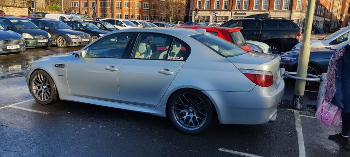 The return of my E60 M5 - Wallet drained - Page 41 - Readers' Cars - PistonHeads UK - The image is a photograph depicting a silver-colored car parked in a parking lot. The car appears to be modified, featuring a prominent rear wing and custom wheel design. It has a sunroof and the bodywork includes additional side cladding. The background shows other vehicles and what seems to be a commercial building with signs that are not legible due to the resolution of the image. There is no visible text providing context or information about the car or its surroundings.