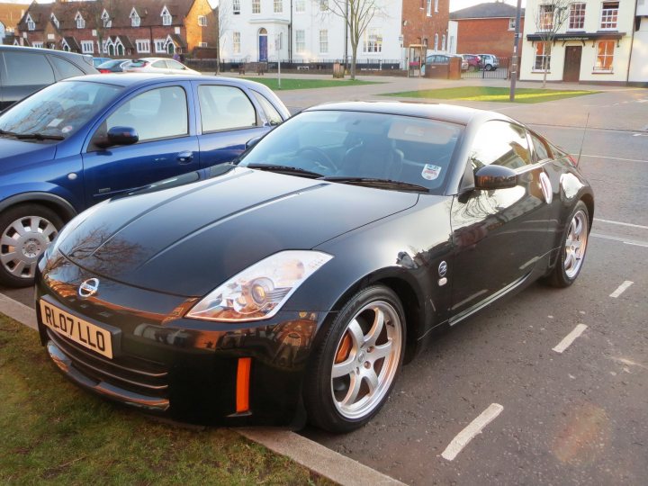 350/370z - Page 1 - Jap Chat - PistonHeads - The image depicts a black sports car parked on a city street. There is another blue car visible in the background, but it is out of focus. The street is bordered by typical urban architecture, with a building on the left side of the photo. A small tree is also visible near the blue car, adding a touch of greenery to the urban setting. The photo appears to be taken during the day, under clear skies.