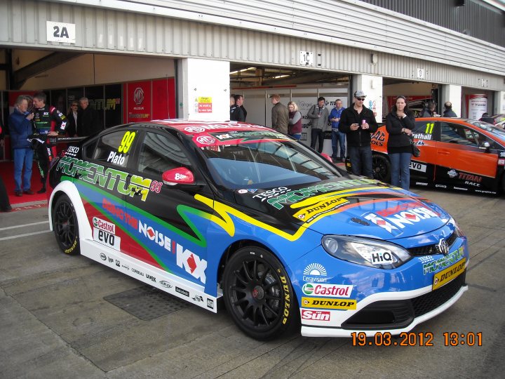 BTCC Media Day. MG6 related - Page 1 - MG - PistonHeads - The image features a vibrant and colorful racing car on a track. The car is prominently displayed in the foreground, adorned with various logos and sponsors' names. The background reveals a bustling scene with a crowd of spectators and other race cars. The photo appears to be taken on a race day, possibly during a pit stop. There's a timestamp on the image indicating it was captured on March 17, 2012 at 13:01.