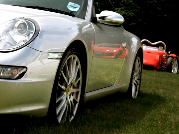 Pistonheads Manor Motors - The image depicts a tranquil scene in a grassy area. A silver convertible car is parked, reflecting the surroundings in its shiny exterior. The reflections on the car's surface add a mirror-like quality to the image. In the distance, there's another car, also parked, which contrasts with the converted car due to its smaller size and red/orange color. The background is serene with trees providing a natural setting. The image exudes a sense of calmness and the beauty of a day in a park.