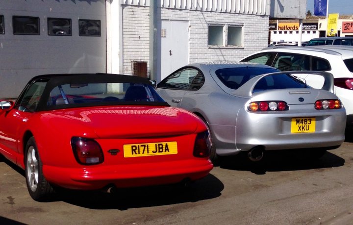 Random TVR pics - Page 399 - General TVR Stuff & Gossip - PistonHeads - The image shows a street scene with three cars parked in front of a white brick building. The most prominent car is a bright red sports car with the license plate "R17 JBA". Next to it is a silver sports car with a license plate that reads "MX89 KXU". The third car, partially visible, is a white vehicle with a license plate starting with "MT63". The scene suggests a casual, day-to-day moment in an urban setting.