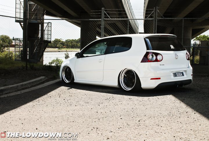 Pictures of decently Modified cars [Vol. 2] - Page 192 - General Gassing - PistonHeads - The image showcases a white two-door car, which appears to be a VW GTI, parked in an outdoor setting. The car is positioned as if it's driving off a parking lot ramp near a bridge or underpass guard rail. The pavement has a slight discoloration or shadow from the bridge's structure, adding depth to the photo. There's a fence and some greenery in the background, providing context for the location. The car has a noticeable modification - an aftermarket exhaust, which adds a sporty touch to its appearance.