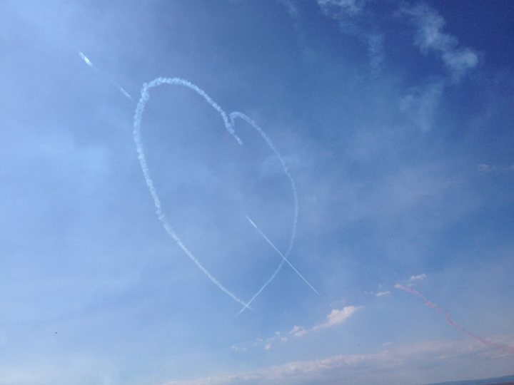 A kite is flying high in the sky - Pistonheads - The image is a photograph capturing a dynamic scene in the clear daytime sky. Two trails of condensed vapor form the configuration of a heart, with one trail forming the left and the other the right side of the heart shape. The sky is a serene blue, with a few clouds scattered in the background. Scattered across the sky, there are a few small yellow objects, possibly other planes or a different type of flying object. The image conveys a sense of motion and the fleeting nature of the contrails.