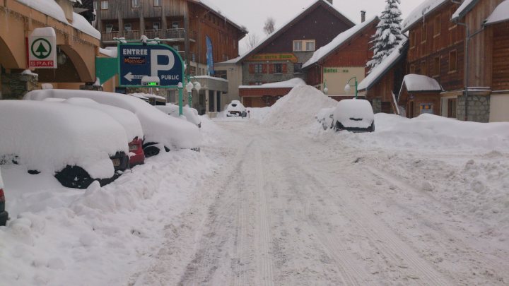 Winter tyres. - Page 1 - Off Road - PistonHeads - The image shows a snow-covered street with residential and commercial buildings lining the sides. The ground is covered in a thick layer of snow, partially filled with tire tracks. The ground-level view emphasizes the heavy snowfall. There is a parking sign visible on the street. Cars are parked along the street and are also covered in snow, highlighting the harsh winter conditions in the city.