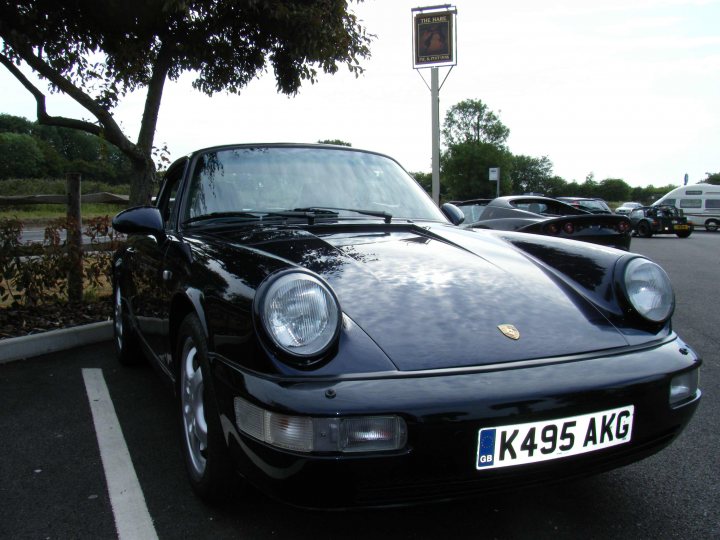 Breakfast Meeting, The Hare, Roxwell 4th June - Page 1 - Kent & Essex - PistonHeads - This image features a black Porsche sports car parked in a parking lot with several other cars. The Porsche is in the foreground, with trees and a sign seen behind it. The license plate of the Porsche is visible with the inscription "K495 AKG." The car has a classic design with round headlights and a smooth, glossy finish. The weather appears to be cloudy.