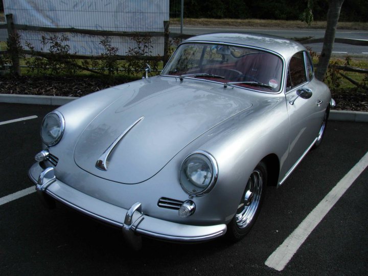 Breakfast Meeting, The Hare, Roxwell 4th June - Page 1 - Kent & Essex - PistonHeads - The image shows a classical silver Porsche 356 parked in a parking lot. The car is facing the viewer, and it appears to be a convertible model. The Porsche is gleaming in the light, reflecting its elegant design. The vehicle is parked in a space designated by white parking lines, indicating an organized parking area. The surrounding area includes a fence with foliage behind it, suggesting a well-maintained environment.