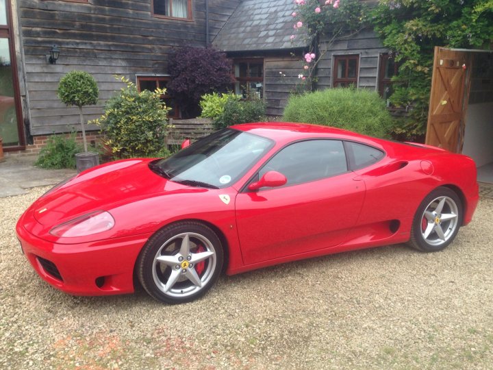 My new toy  - Page 1 - Ferrari V8 - PistonHeads - The image showcases a sporty red Ferrari parked on a gravel driveway. The car's sleek design, particularly the enlarged side vents, is a distinguishing feature. Adjacent to the driveway is a wooden cabin, and in the background, there's a house with a distinctive black roof and pink flowers. The setting suggests a residential area with a blend of modernity and rustic charm.