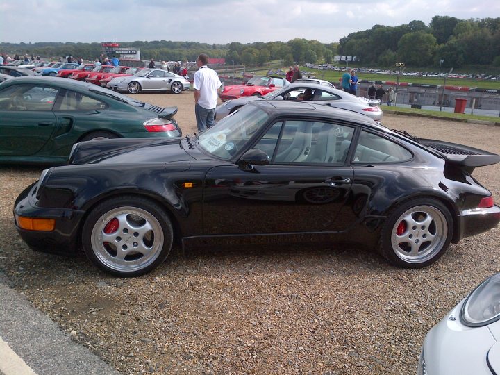 964 turbo 3.6 - Page 1 - Porsche Classics - PistonHeads - The image captures a scene of a gravel parking lot filled with a variety of cars, mainly sports models. These cars are positioned in such a way that they are facing the same direction, suggesting a gathering or event. The background presents a serene landscape with trees and a sky partly cloudy, providing a contrast to the urban atmosphere suggested by the cars in the foreground. The image is taken from a slightly elevated angle, allowing a comprehensive view of the parking lot and the surrounding environment. The variety of car colors and models contributes to the diversity of the scene.