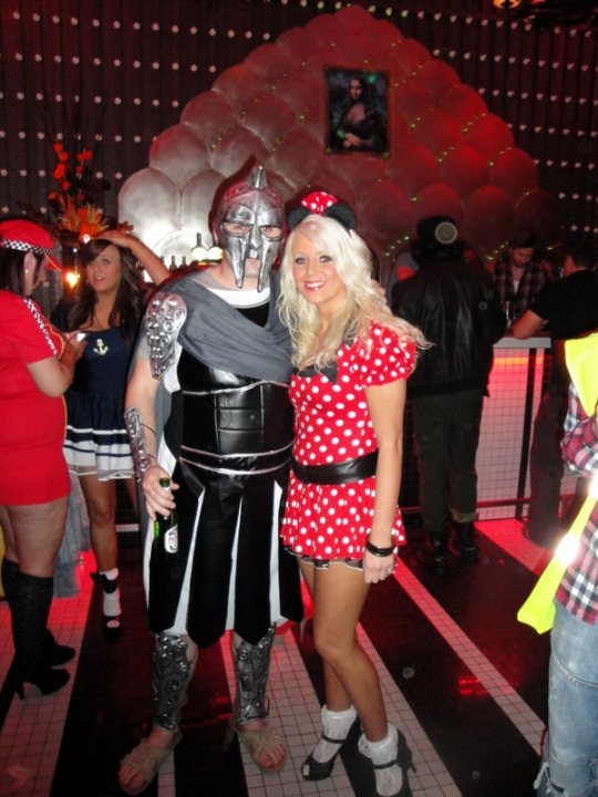 Pistonheads Costume Halloween - The image portrays a lively scene at what seems to be a costume party. A man wearing a silver gladiator helmet and a matching tunic stands beside a woman dressed as Minnie Mouse, complete with her iconic red and white dress. The backdrop of the photograph includes geometric patterns, adding to the festive atmosphere of the party. Other party-goers are visible in the background; a woman in a red dress is holding a glass, presumably filled with a beverage, while another man in a black t-shirt can be seen wearing a matching baseball cap. The faces of all individuals in the picture are turned towards the camera, suggesting they are posing for the photograph. The composition of the image, along with the attire of the people, indicates that the event is a themed party where guests are dressed in colorful and diverse costumes.