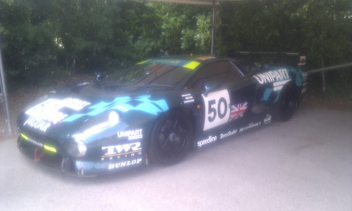 Jaguar XJ220C Le Mans racer at the FOS - did it run ? - Page 1 - Goodwood Events - PistonHeads - This image features a Jaguar racing car parked near a covered area with trees in the background. The car is predominantly black with a striking blue and white XJR racer livery. On the side and back, there are number 50 written with a Union Jack motif, suggesting it is either a number from racing and a British flag. The car's position and the clear sky indicate that it is during the day.