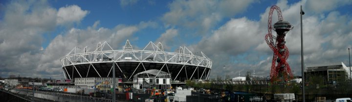 Visit to the Olympic Park - Page 1 - Sports - PistonHeads - The image showcases a modern architectural structure, which appears to be a stadium. Interestingly, the stadium is predominantly black with white accents, providing a striking contrast to the surrounding environment. In the distance, behind the stadium, there's a roller coaster adding an element of fun and excitement. The sky above is a clear blue with numerous white clouds scattered across it, indicating a pleasant day with good weather conditions. The lower part of the image features a construction site, hinting at the continuous development in the area.