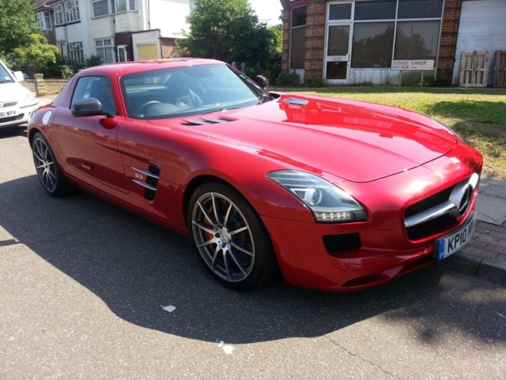 A red car with a surfboard on top of it - Pistonheads - The image showcases a vibrant red Mercedes sports car parked on the sidewalk of a neighborhood street. The car is positioned alongside a white vehicle. The car's glossy paint and shiny chrome details are clearly visible. The vehicle is equipped with large rims and five-spoke wheels that add to its stylish appearance. The car is parked in a small lot next to a building, which has a sign that reads "Safety Gate" in the background. The setting suggests a calm, suburban atmosphere.