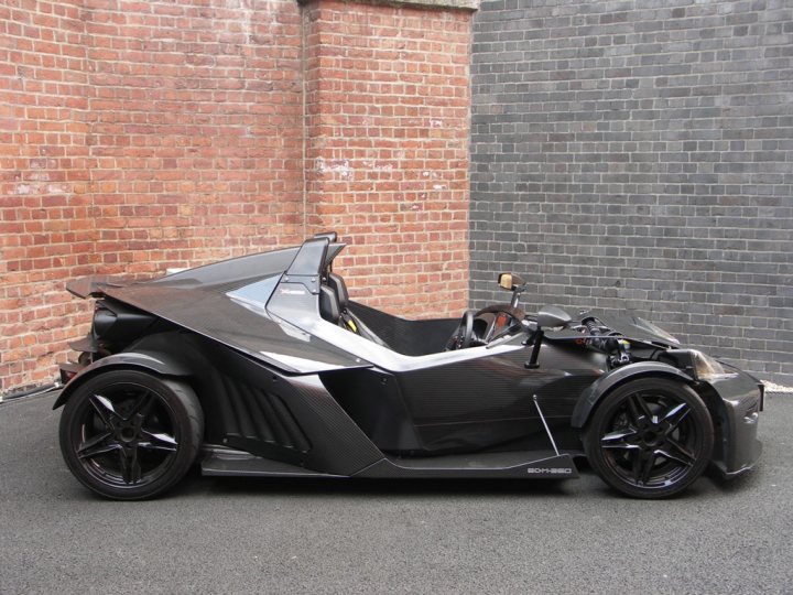 Friday Pistonheads Beastie Picking - In the image, a sleek black sports car is parked amidst an urban setting. The car, which appears to be a Formula Race Car, is positioned against a contrasting brick wall and is unique in its design with a front-wheel drive feature. The vehicle is dark, possibly black, which matches the monochromatic brick wall in the background. The car's design suggests high performance capabilities, and it's placed in such a way that it's facing towards the left side of the image.