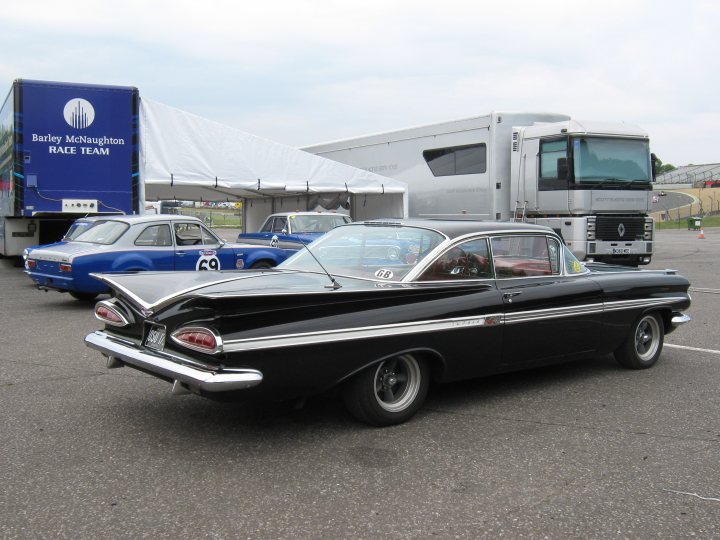 Lets see a picture of your classic(s) - Page 269 - Classic Cars and Yesterday's Heroes - PistonHeads - This image features a classic black convertible coupe car with a vintage design, parked in front of a pale green sports coach bus. The car is the centerpiece of the image, placed on the blacktop of an asphalt parking lot. In the background, there are other cars parked, including a blue vintage car partially visible. The sky above is overcast, suggesting an outdoor event or gathering might be taking place. There is a white tent structure visible in the background, providing shelter and possibly serving as a VIP or event area. The setting appears to be a race rally or a classic car show, indicated by the text "Barley McNaght Race Team" on a flag-like structure.