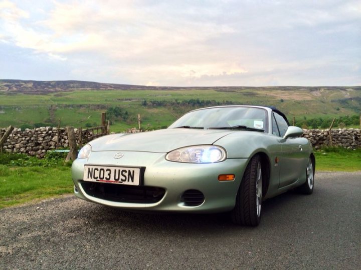 my mx5 - Page 1 - Mazda MX5/Eunos/Miata - PistonHeads - The image showcases a silver-colored Mazda sports car, clearly distinguished by its NCO3 USN license plate, parked along the side of a road. The car is set against the backdrop of a verdant green hillside dotted with patches of sunlight. A stone wall and what appears to be a wooden post provide additional context to the scene. The play of light on the car suggests it might be late afternoon or early morning.