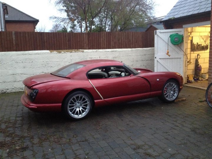 Just a little vid of my cerbera - Page 1 - Cerbera - PistonHeads - The image features a vibrant red, two-door sports car with a smoked grey tint parked on a brick driveway. The car has a sleek design with black accents and a large spoiler on the rear. The car's large wheels have silver rims and are covered in red dirt, suggesting it has been driven on rough terrain. To the right of the car, there is an open garage with a green gas pump. Closer to the garage, a regular bulldog is visible, likely a pet, adding a touch of homeliness to the scene.