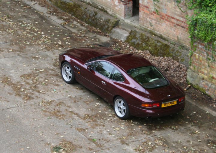 Your DB7 pictures. - Page 1 - Aston Martin - PistonHeads - The image features a dark red sports car parked on a concrete surface that appears slightly damp, possibly from recent rain. Leaves are scattered around, suggesting that autumn has recently passed. The car exhibits a sleek and streamlined design, with its fixed rear spoiler, indicative of high-performance racing cars. It's positioned on a slight incline, adding a dynamic element to the setting. The background includes a brick wall and a pathway, contributing to an urban or suburban feel.