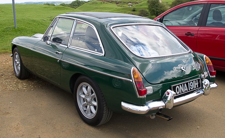 Looking for a MGC GT . . .  - Page 1 - MG - PistonHeads - The image shows a vintage green car parked on a gravel surface next to a red car. The green car has a hood ornament in the middle of the front hood, and it also features an analog clock-like instrument panel on the dashboard. The car's vintage design and its bright green color, as well as the license plate plate that starts with "OMA" and "19H," indicate that this is not a common contemporary vehicle. In the background, there is a landscape that includes grass, trees, and a blue sky, suggesting a peaceful, possibly rural setting.