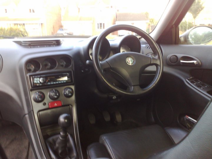 Romeo White Pistonheads Polar Alfa - The image presents the interior of a silver car, viewed from the rear passenger seat. The cockpit area is prominently visible, featuring a steering wheel centered in the frame, control dials in the center console, which appear to be a mix of buttons and dials, mimicking a manual transmission layout, and an air vent above it. To the right of the wheel, there is a gear shift lever, indicating a hybrid or manual transmission car. There are no visible people in the image. The image has a realistic color representation and is taken during daytime, as suggested by the bright lighting in the scene.